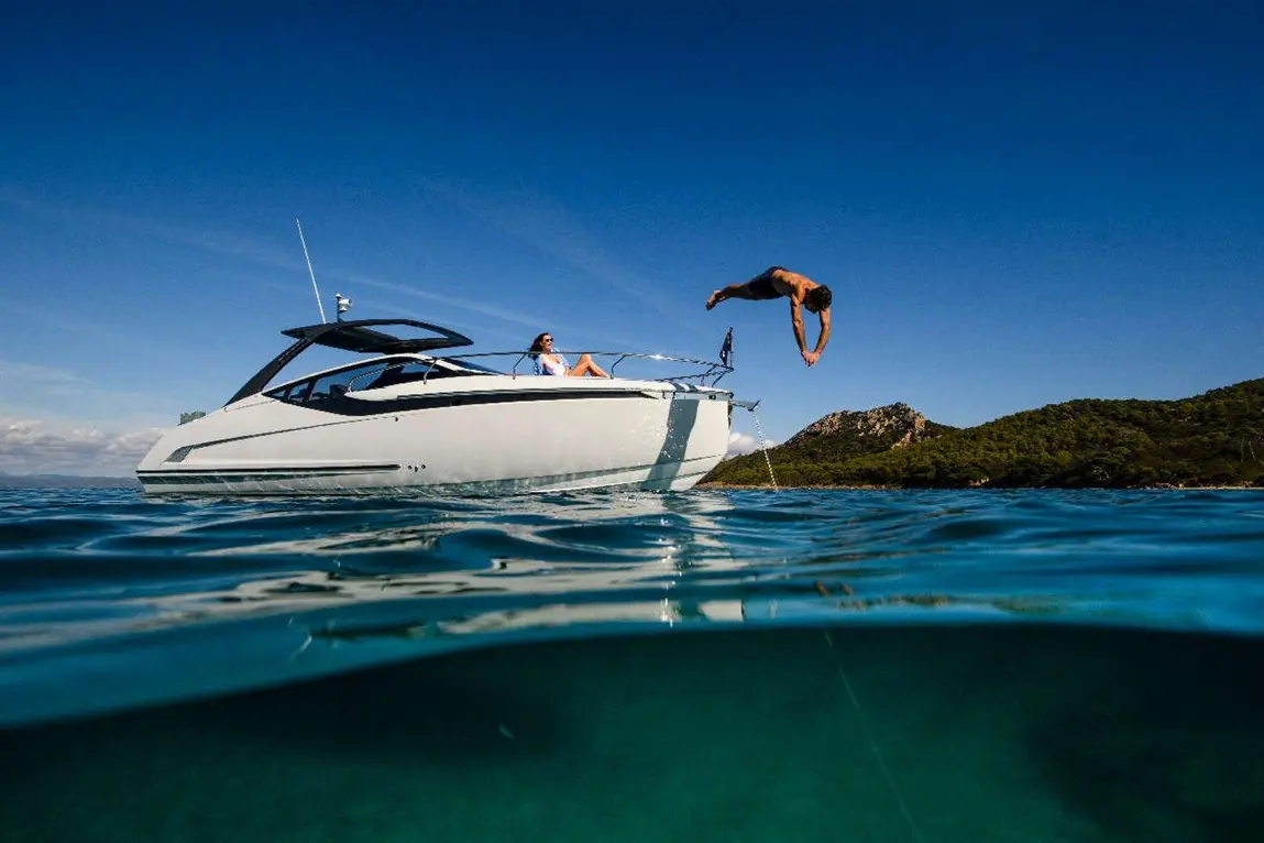 A man dives off a luxury motorboat into clear turquoise water, while a woman relaxes on the boat under a blue sky.